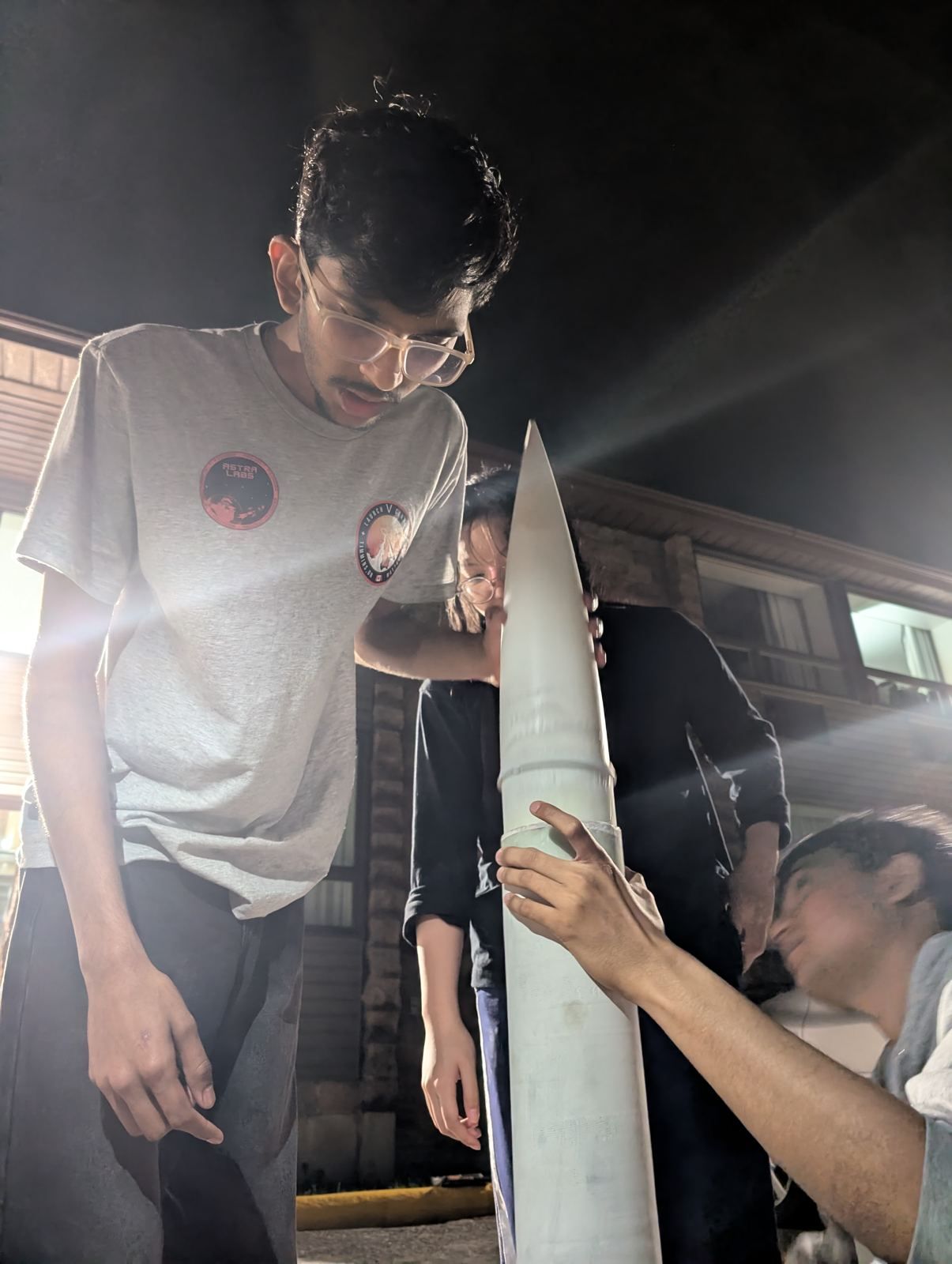 A team member under work lights at night checking the nose of a rocket held upright